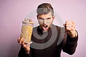 Young blond man with beard and blue eyes holding bottle of Italian dry pasta macaroni annoyed and frustrated shouting with anger,