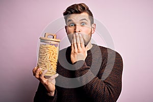 Young blond man with beard and blue eyes holding bottle of Italian dry pasta macaroni cover mouth with hand shocked with shame for