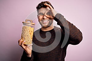 Young blond man with beard and blue eyes holding bottle of Italian dry pasta macaroni with happy face smiling doing ok sign with