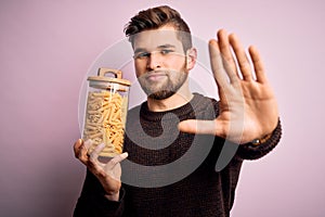 Young blond man with beard and blue eyes holding bottle of Italian dry pasta macaroni with open hand doing stop sign with serious