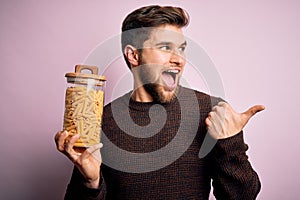 Young blond man with beard and blue eyes holding bottle of Italian dry pasta macaroni pointing and showing with thumb up to the