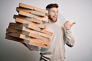 Young blond man with beard and blue eyes holding cardboards of pizza over white background pointing and showing with thumb up to