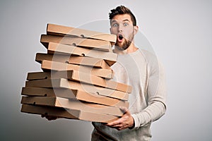 Young blond man with beard and blue eyes holding cardboards of pizza over white background scared in shock with a surprise face,