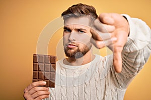 Young blond man with beard and blue eyes holding chocolate bar over yellow background with angry face, negative sign showing