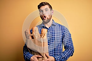 Young blond man with beard and blue eyes holding paper bag with bread scared in shock with a surprise face, afraid and excited