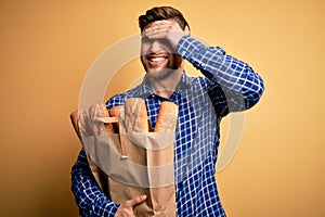 Young blond man with beard and blue eyes holding paper bag with bread stressed with hand on head, shocked with shame and surprise