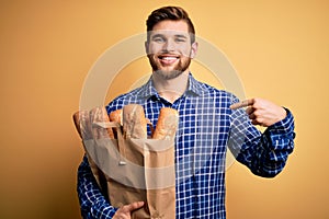Young blond man with beard and blue eyes holding paper bag with bread with surprise face pointing finger to himself