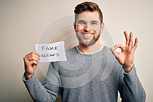 Young blond man with beard and blue eyes holding paper with fake news message doing ok sign with fingers, excellent symbol