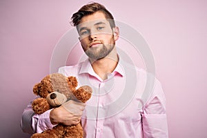 Young blond man with beard and blue eyes holding teddy bear toy over pink background with a confident expression on smart face