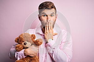 Young blond man with beard and blue eyes holding teddy bear toy over pink background cover mouth with hand shocked with shame for