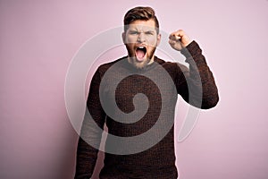 Young blond man with beard and blue eyes wearing casual sweater over pink background angry and mad raising fist frustrated and