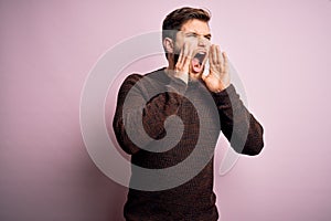 Young blond man with beard and blue eyes wearing casual sweater over pink background Shouting angry out loud with hands over mouth