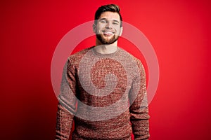 Young blond man with beard and blue eyes wearing casual sweater over red background with a happy and cool smile on face