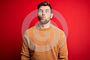 Young blond man with beard and blue eyes wearing casual sweater over red background making fish face with lips, crazy and comical