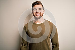 Young blond man with beard and blue eyes wearing green sweater over white background with a happy and cool smile on face