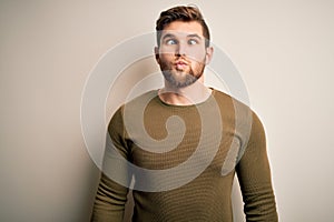 Young blond man with beard and blue eyes wearing green sweater over white background making fish face with lips, crazy and comical