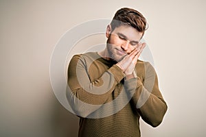 Young blond man with beard and blue eyes wearing green sweater over white background sleeping tired dreaming and posing with hands