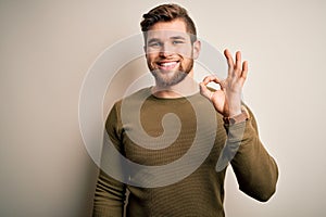 Young blond man with beard and blue eyes wearing green sweater over white background smiling positive doing ok sign with hand and