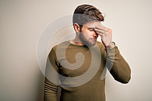 Young blond man with beard and blue eyes wearing green sweater over white background tired rubbing nose and eyes feeling fatigue