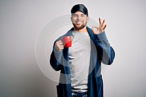 Young blond man with beard and blue eyes wearing pajama drinking cup of coffee doing ok sign with fingers, excellent symbol