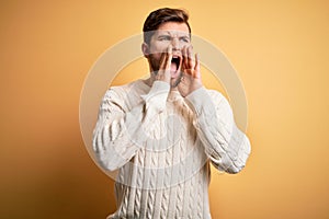 Young blond man with beard and blue eyes wearing white sweater over yellow background Shouting angry out loud with hands over