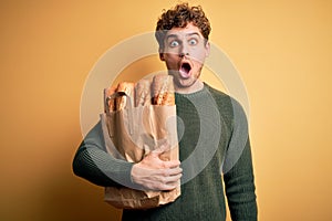 Young blond man with curly hair holding paper bag with bread over yellow background scared in shock with a surprise face, afraid