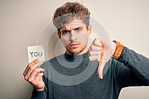 Young blond man with curly hair holding paper with you message over white background with angry face, negative sign showing