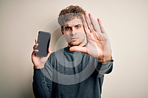 Young blond man with curly hair showing smartphone standing over isolated white background with open hand doing stop sign with