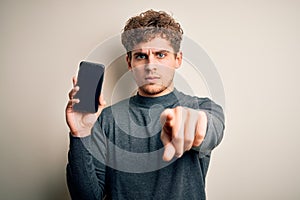 Young blond man with curly hair showing smartphone standing over isolated white background pointing with finger to the camera and