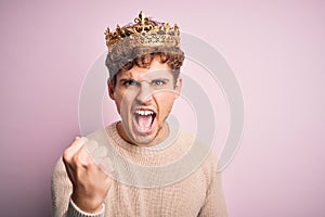 Young blond man with curly hair wearing golden crown of king over pink background angry and mad raising fist frustrated and