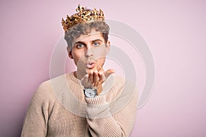 Young blond man with curly hair wearing golden crown of king over pink background looking at the camera blowing a kiss with hand