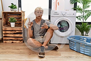 Young blond man doing laundry using smartphone pointing up looking sad and upset, indicating direction with fingers, unhappy and