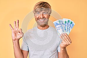 Young blond man holding south african rand banknotes doing ok sign with fingers, smiling friendly gesturing excellent symbol