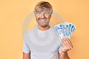 Young blond man holding south african rand banknotes looking positive and happy standing and smiling with a confident smile