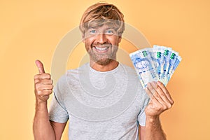 Young blond man holding south african rand banknotes smiling happy and positive, thumb up doing excellent and approval sign