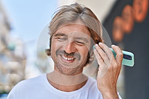 Young blond man miling confident listening audio message by the smartphone at street