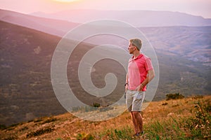 A young blond man in a pink shirt conquered the top of the mountain at sunset. Beautiful mountain landscape with a gradient. The c