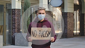 A young and blond man protester activist is wearing a medical mask holds a cardboard poster with the slogan NO RACISM. A