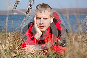 Young blond man in red jacket sit on the seashore.
