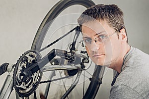 Young blond man with short haircut in a gray t-shirt with glasses sitting near spinning wheel of dirty broken bicycle