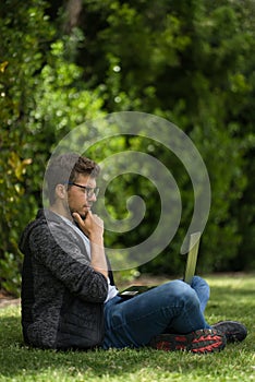 A young blond man is sitting on the grass of a park with his laptop. He is with her legs crossed and he seems to be thinking about
