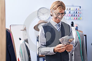 Young blond man tailor smiling confident holding dollars at clothing factory