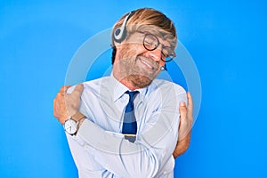 Young blond man wearing call center agent headset hugging oneself happy and positive, smiling confident