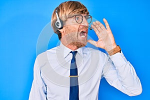 Young blond man wearing call center agent headset shouting and screaming loud to side with hand on mouth