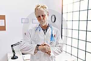 Young blond man wearing doctor uniform and stethoscope at clinic checking the time on wrist watch, relaxed and confident
