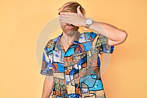 Young blond man wearing summer shirt covering eyes with hand, looking serious and sad