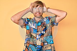 Young blond man wearing summer shirt crazy and scared with hands on head, afraid and surprised of shock with open mouth