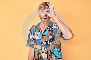 Young blond man wearing summer shirt doing ok gesture shocked with surprised face, eye looking through fingers