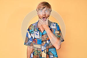 Young blond man wearing summer shirt looking fascinated with disbelief, surprise and amazed expression with hands on chin