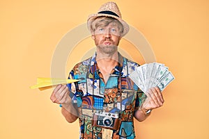 Young blond man wearing summer style holding paper plane and dollars puffing cheeks with funny face